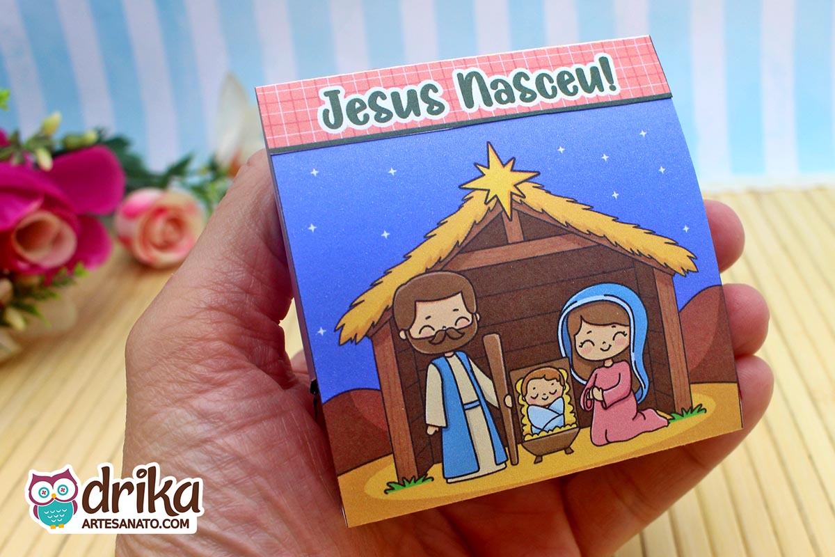 Mão segurando porta-bombom "Jesus Nasceu" com ilustração da Sagrada Família e céu estrelado, mostrando o tamanho e design.