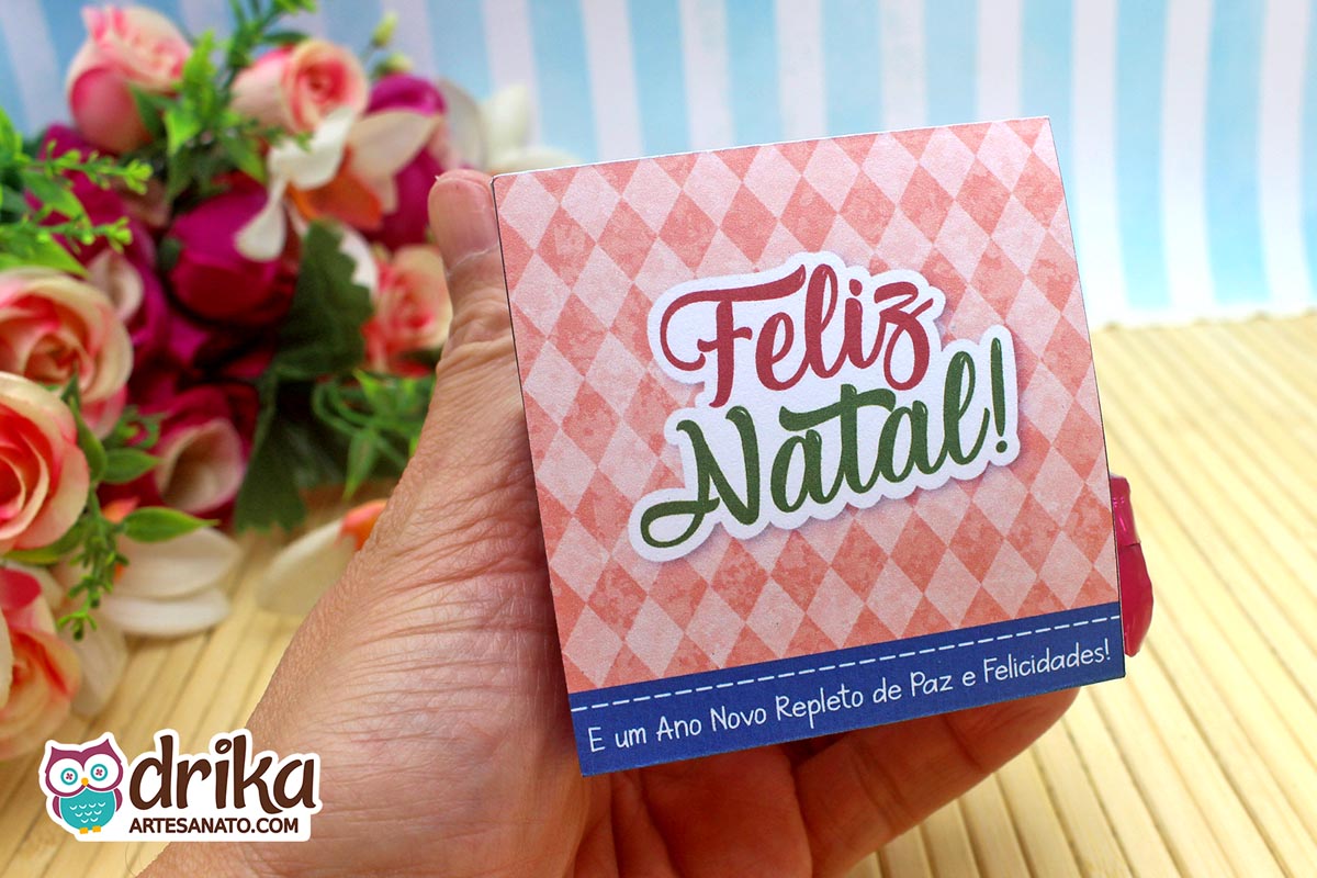 Mão segurando a parte de trás do porta-bombom "Feliz Natal" com xadrez rosa e mensagem de Ano Novo em fundo azul.