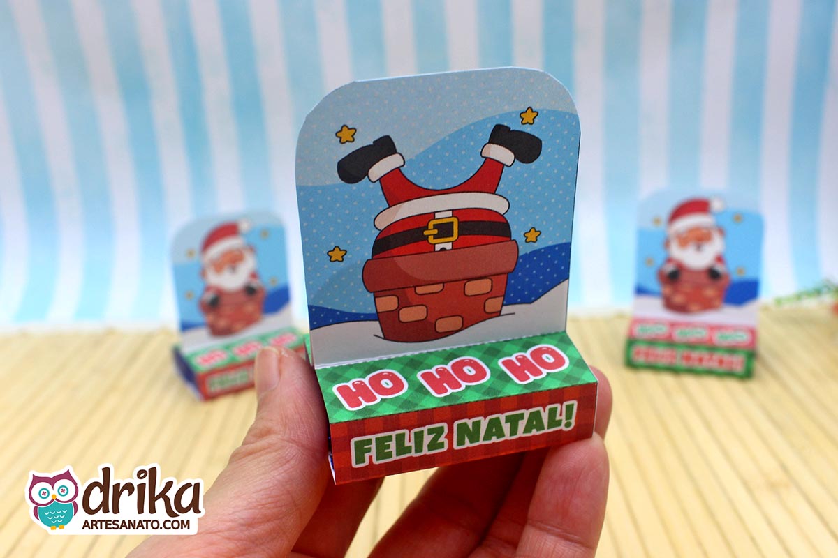 Mão segurando porta-bis duplo de papel com Papai Noel de pernas para o ar em chaminé, com base verde e estrelas