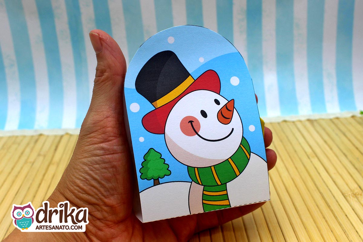 Mão segurando porta-bombom de boneco de neve de papel, branco com chapéu preto e vermelho, cachecol verde e amarelo, nariz de cenoura