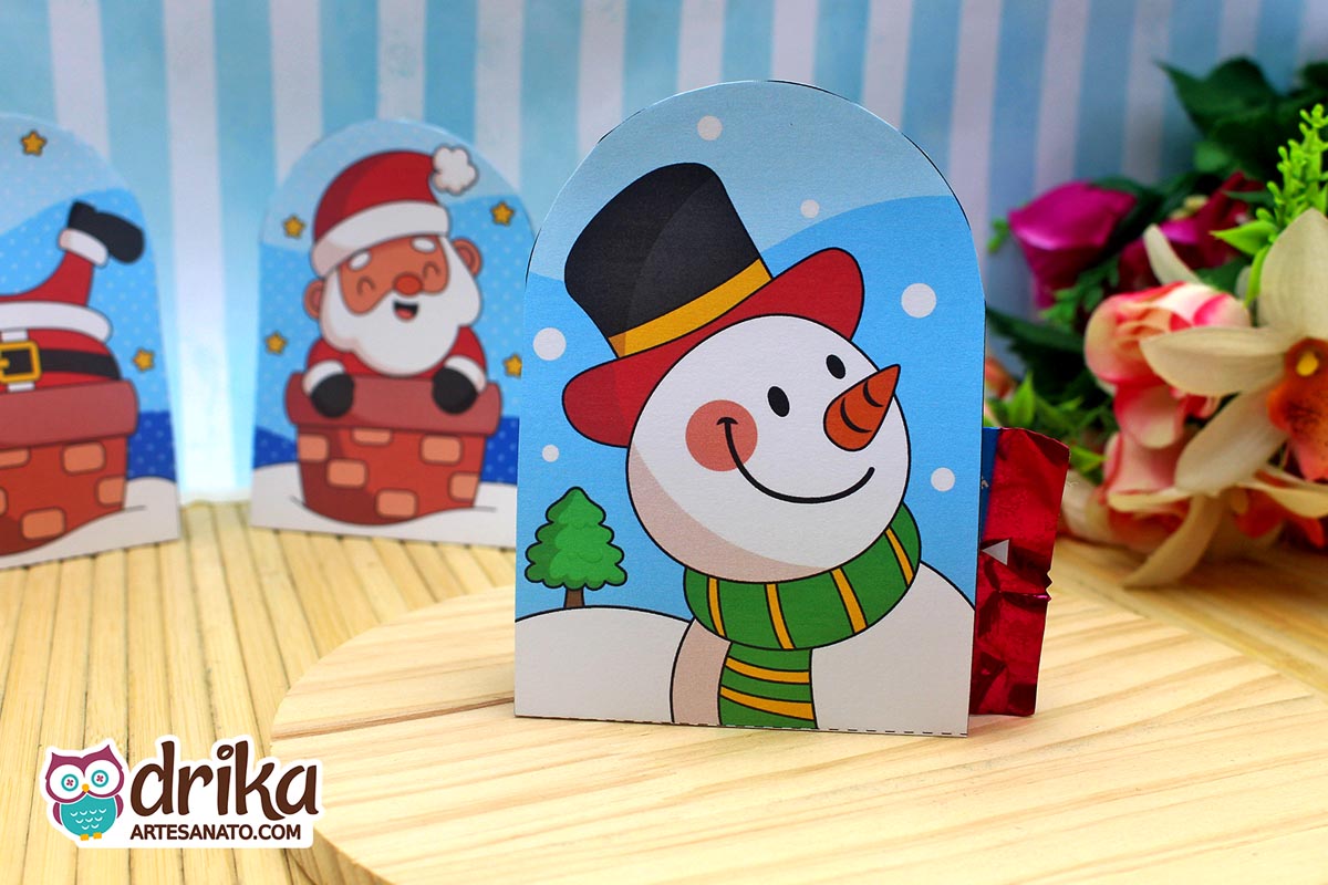 Porta-bombom de boneco de neve de papel com bombom vermelho saindo da lateral, sobre base de madeira redonda
