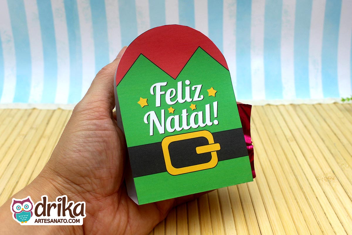 Mão segurando um porta-bombom de papel verde com tema de elfo, cinto preto e fivela dourada, escrito "Feliz Natal"