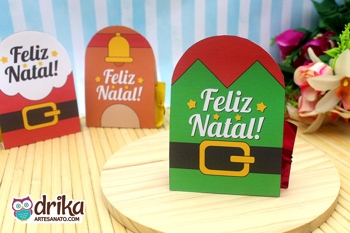 Três porta-bombons de Natal de papel em uma base de madeira, com temas de elfo, Papai Noel e rena, todos com "Feliz Natal"