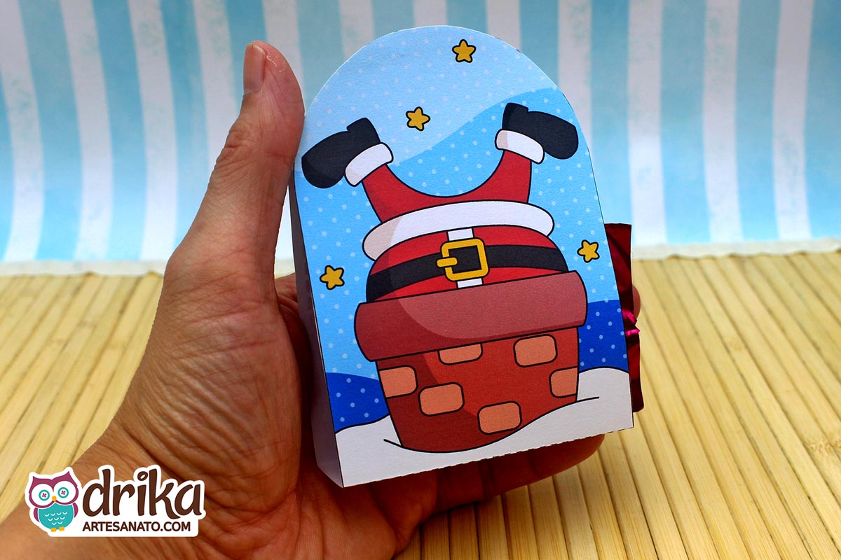 Mão segurando porta-bombom de papel com Papai Noel de pernas para cima descendo por chaminé de tijolos vermelhos, céu azul com estrelas