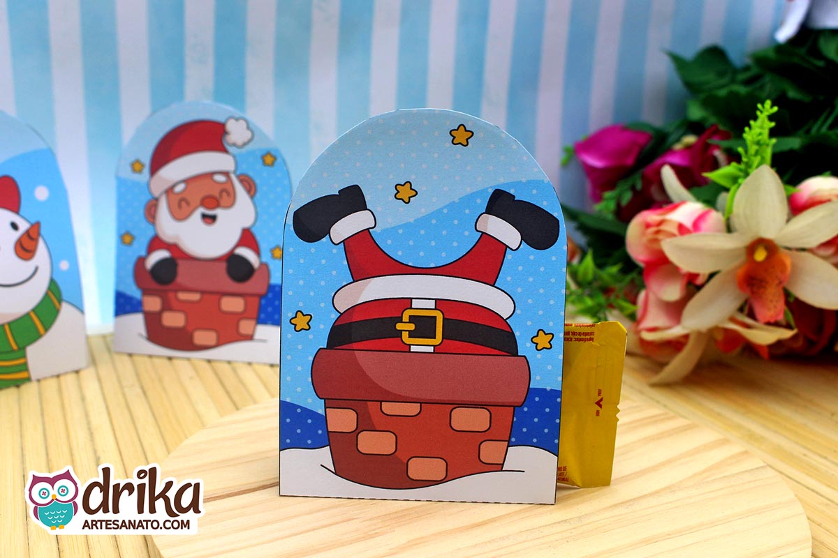 Porta-bombom de papel com Papai Noel de pernas para cima na chaminé, em mesa de madeira clara com outros modelos desfocados
