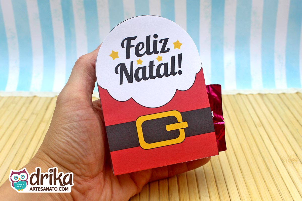 Mão segurando porta-bombom de papel com roupa de Papai Noel vermelha, cinto preto e fivela dourada, e "Feliz Natal" em nuvem branca