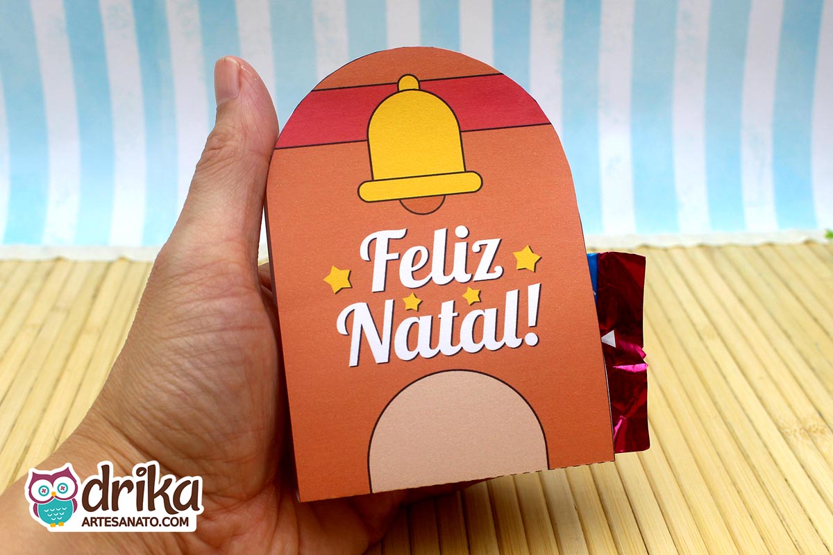 Mão segurando um porta-bombom de rena marrom com sininho dourado e a frase "Feliz Natal!" com estrelinhas