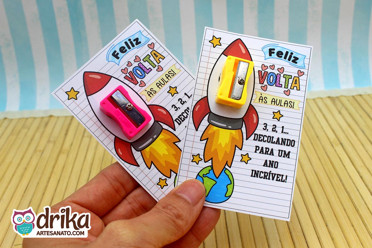 Mão segurando dois cartões de Volta às Aulas com foguetes, um com apontador rosa e outro com apontador amarelo encaixados