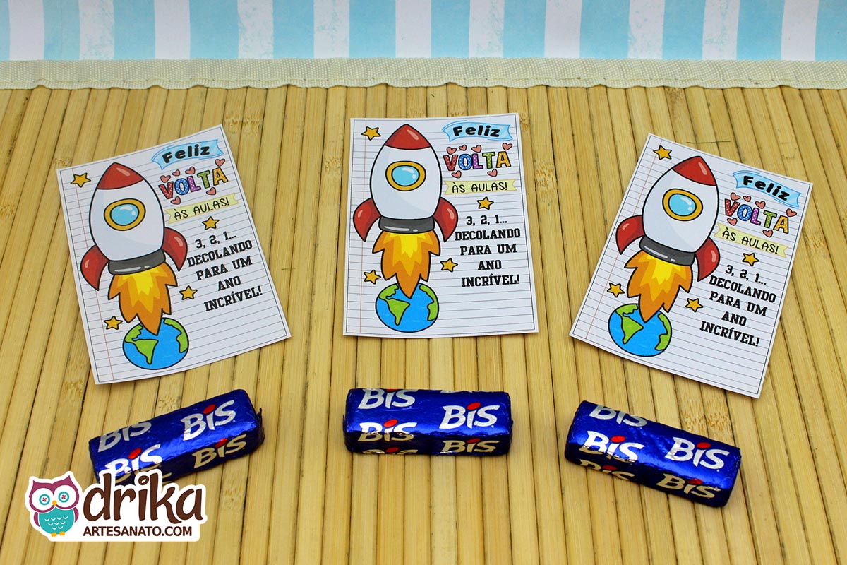Três cartões porta-bis de foguete com tema Volta às Aulas e três chocolates Bis azuis sobre esteira de bambu