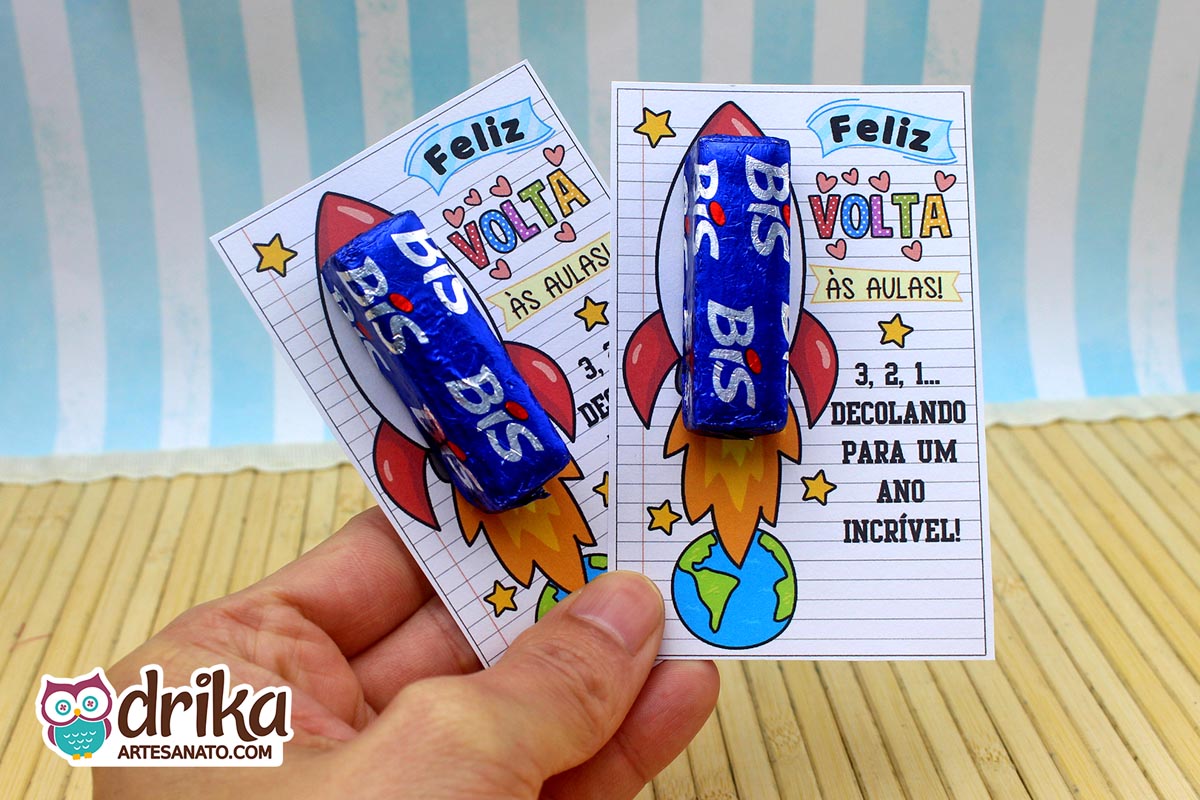 Mão segurando dois cartões porta-bis de foguete com chocolates Bis azuis encaixados, prontos para a Volta às Aulas