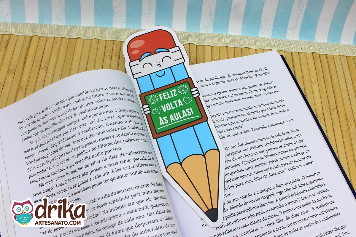 Cartão porta-lápis gigante azul com rosto feliz e lousa "Feliz Volta às Aulas" usado como marca-páginas em livro aberto