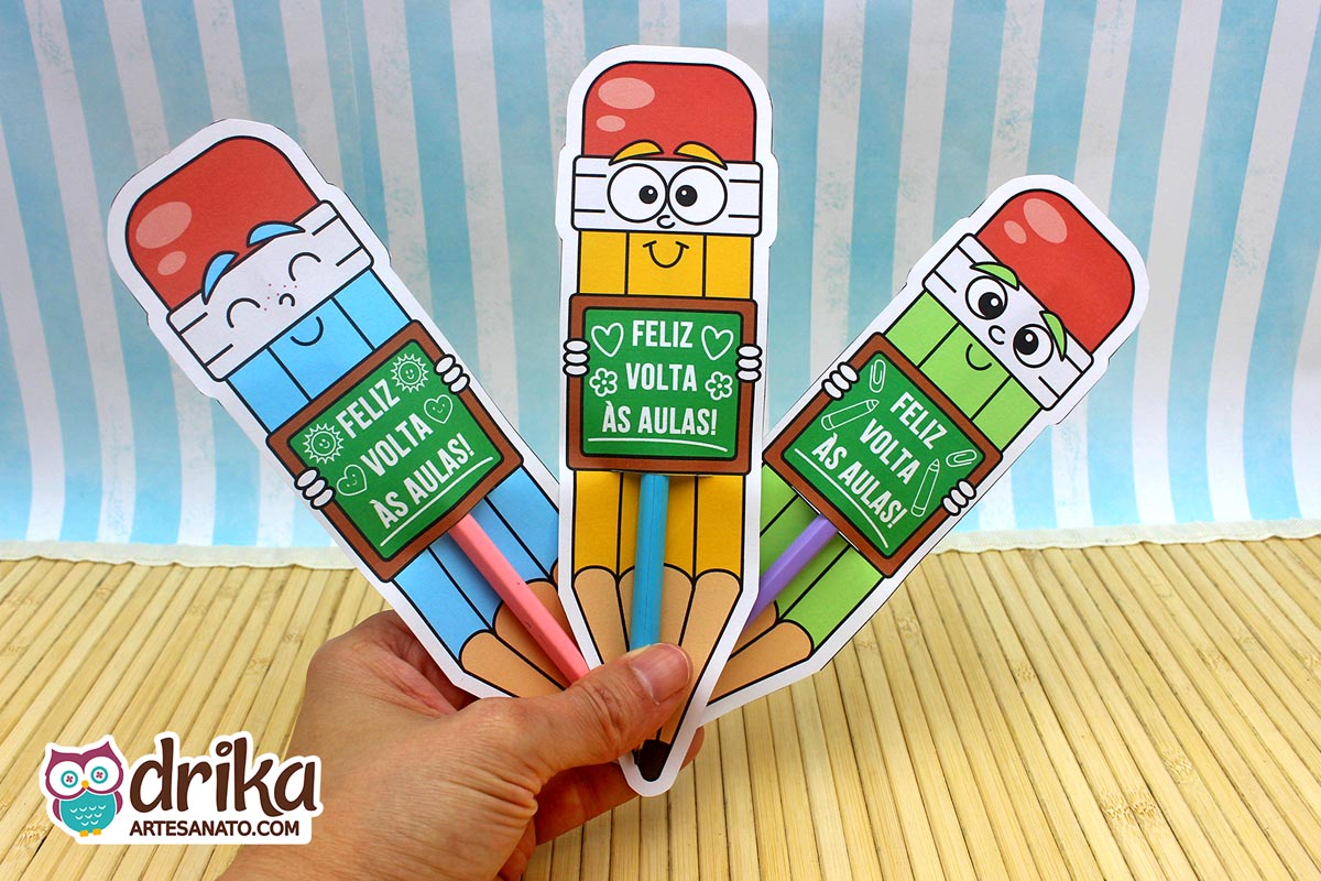 Mão segurando três cartões porta-lápis gigantes de meninos (azul, amarelo, verde) com lápis de cor inseridos
