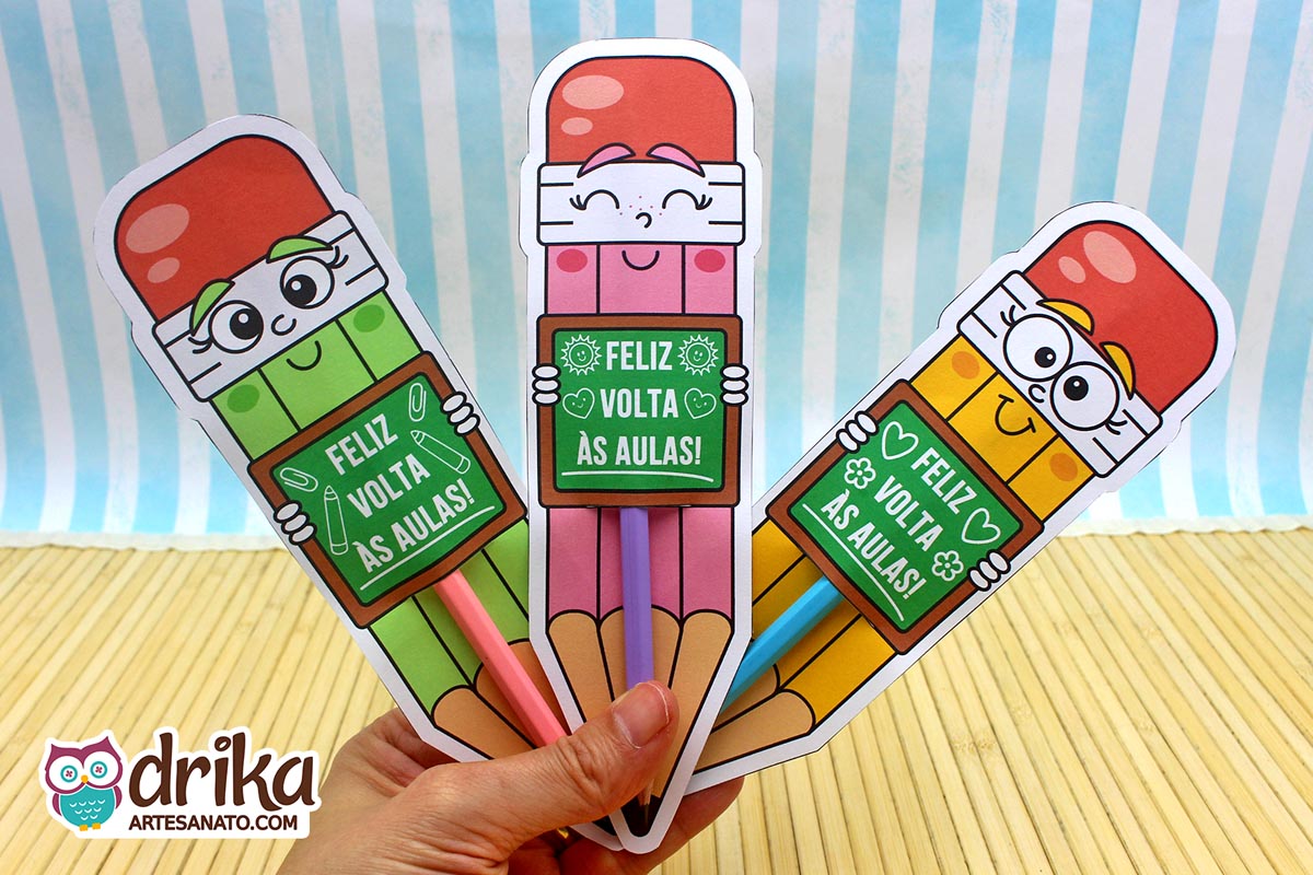 Mão segurando três cartões porta-lápis gigantes de meninas, nas cores amarelo, rosa e verde, com lápis de verdade