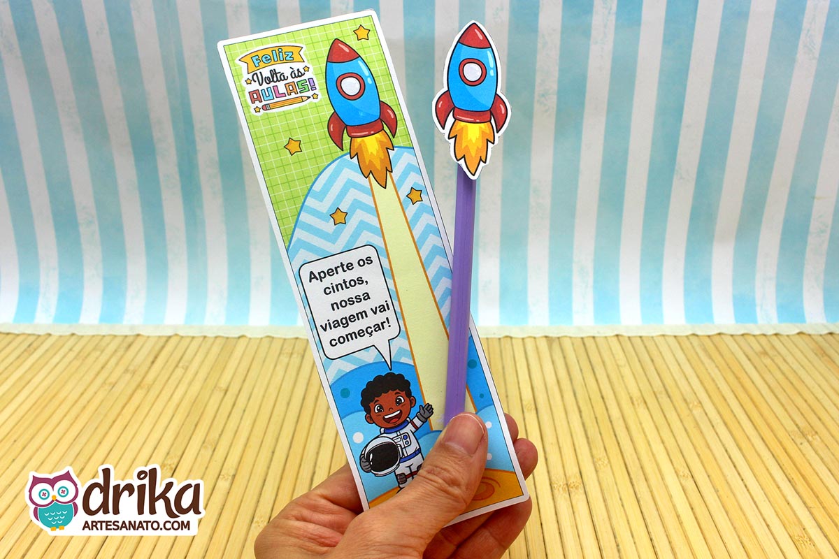 Mão segurando cartão porta-lápis de astronauta menino de cabelo crespo, com lápis roxo e foguete 3D em destaque