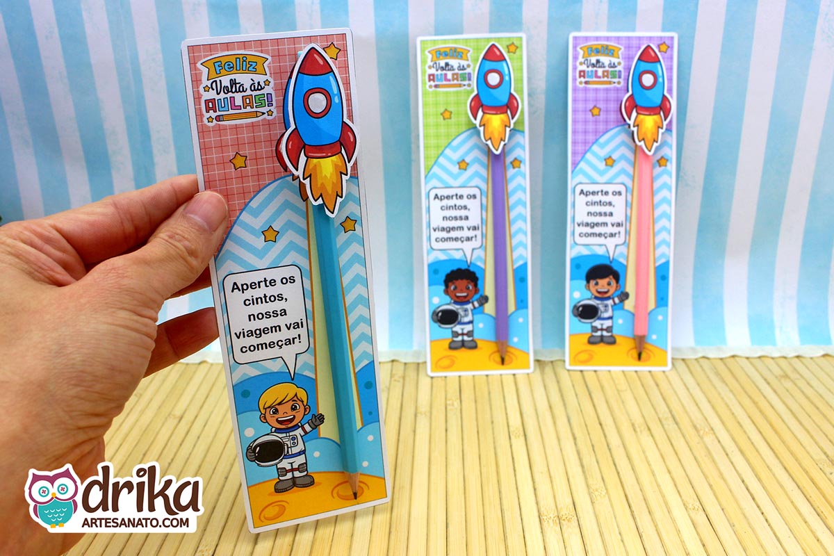 Mão segurando cartão porta-lápis de astronauta menino loiro, com lápis azul e foguete 3D, e outros dois cartões desfocados