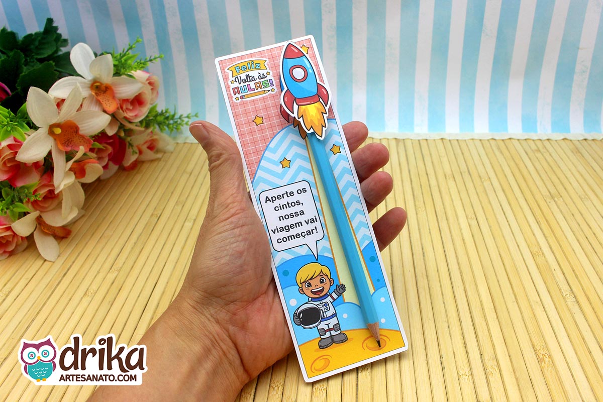 Mão segurando cartão porta-lápis com astronauta menino loiro, lápis azul e foguete 3D, sobre fundo de bambu