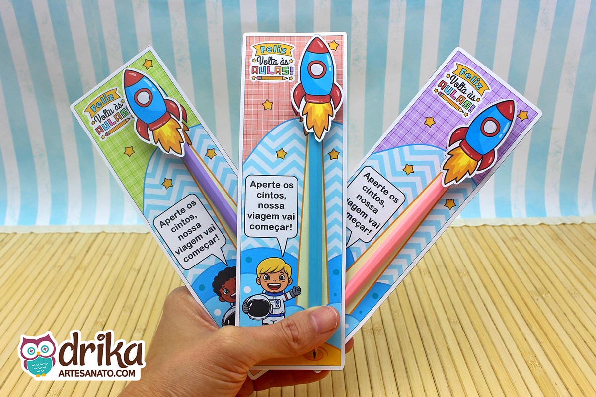 Mão segurando três cartões porta-lápis de astronautas meninos com foguetes 3D nos lápis e a frase "Aperte os cintos, nossa viagem vai começar"