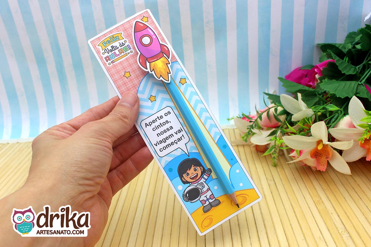 Mão segurando cartão porta-lápis verde com menina astronauta de cabelo preto liso, lápis azul e foguete rosa