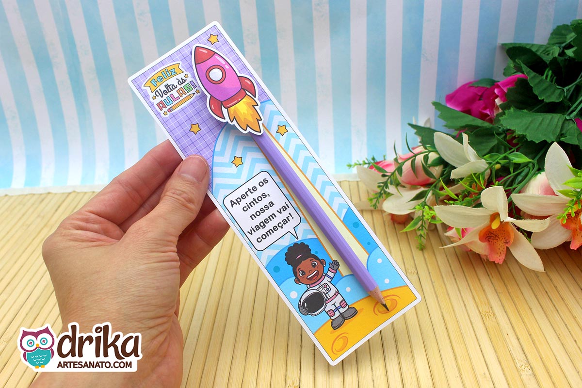 Mão segurando cartão porta-lápis roxo com menina astronauta de cabelo crespo, lápis roxo e foguete rosa