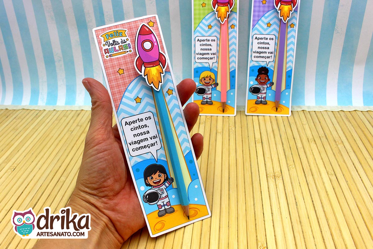 Mão segurando cartão porta-lápis de menina astronauta de cabelo preto liso com foguete e lápis azul