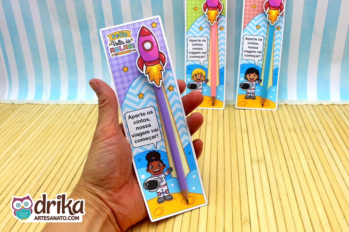 Mão segura cartão porta-lápis roxo com menina astronauta de cabelo crespo, lápis lilás e foguete rosa 3D. Fundo quadriculado e estrelas