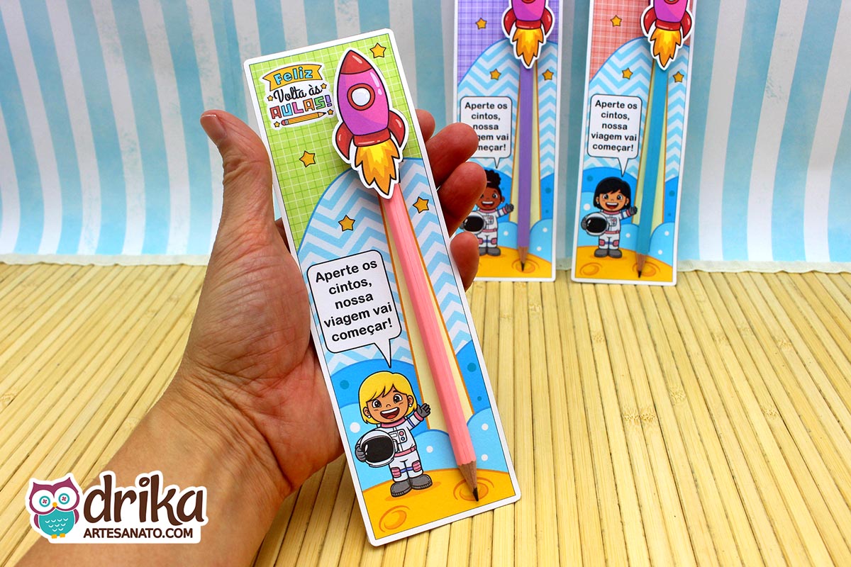 Mão segura cartão porta-lápis verde com menina astronauta loira, lápis rosa e foguete rosa 3D. Fundo quadriculado e estrelas