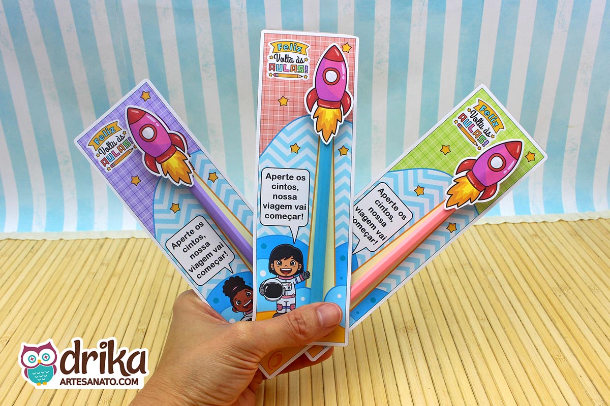 Mão segurando três cartões porta-lápis de astronautas meninas, com lápis coloridos e foguetes 3D.