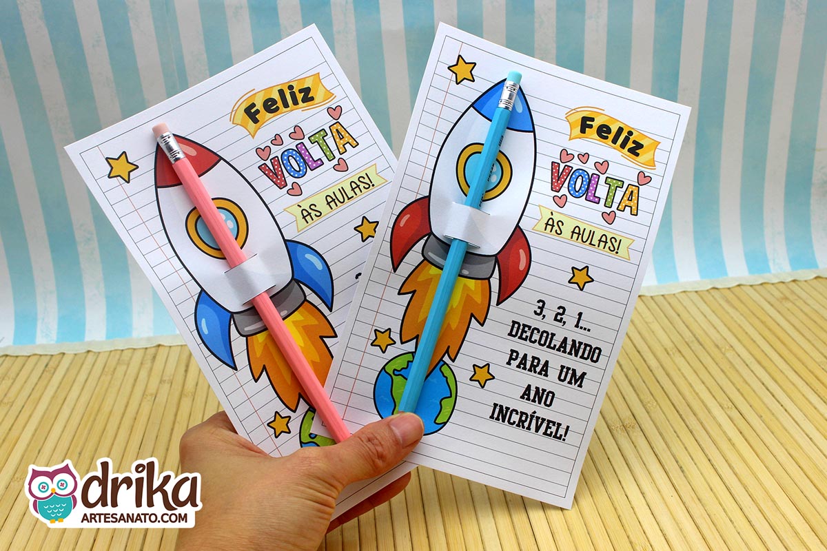 Mão segurando dois cartões porta-lápis de foguete, um com lápis rosa e outro com lápis azul encaixados