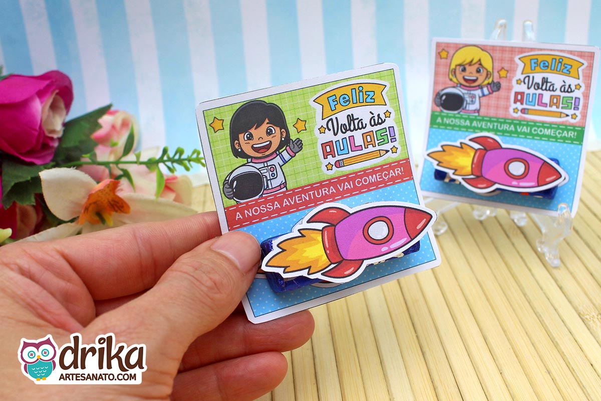 Mão segurando cartão porta-bis com menina astronauta de cabelo preto liso, fundo verde quadriculado e foguete 3D rosa no bis