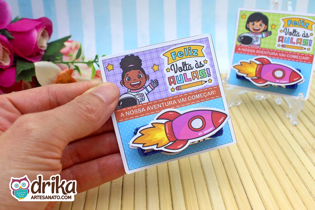 Mão segurando cartão porta-bis com menina astronauta de cabelo crespo, fundo roxo quadriculado e foguete 3D rosa no bis