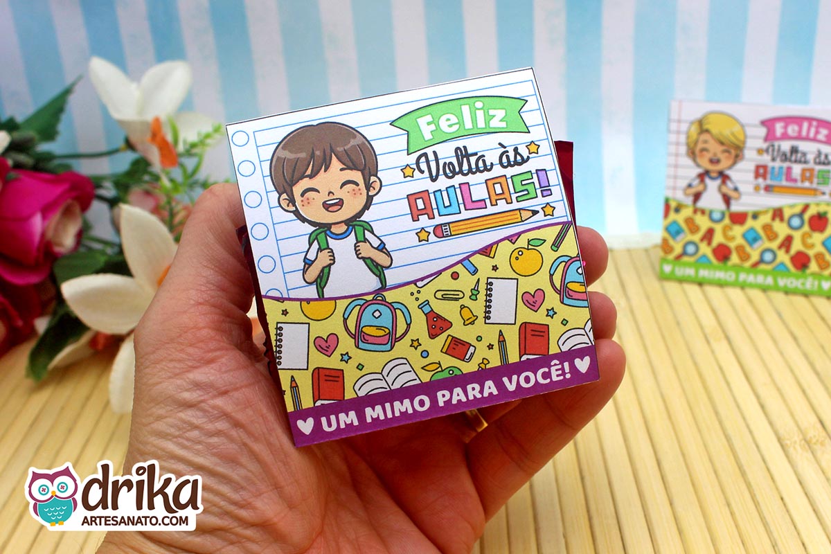 Mão segurando porta-bombom de papel com menino de cabelo castanho e estampa de mochilas e cadernos em fundo de caderno