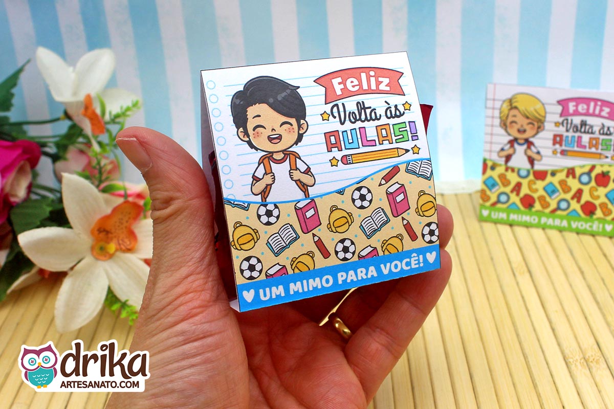 Mão segurando porta-bombom de papel com menino de cabelo preto liso, mochila e frase "Feliz Volta às Aulas"