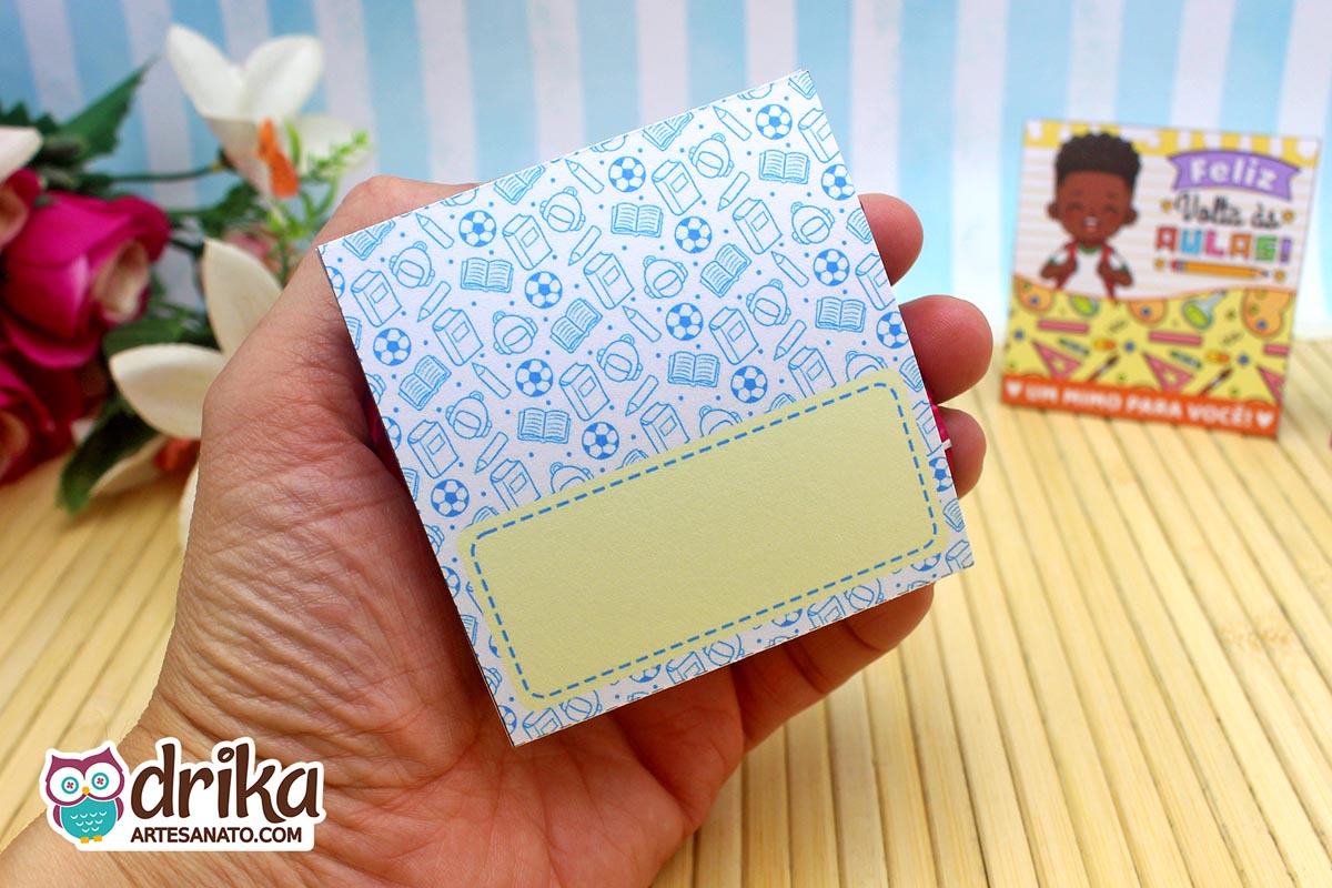 Mão segurando o verso de porta-bombom de papel com padrão de material escolar azul claro e espaço para nome