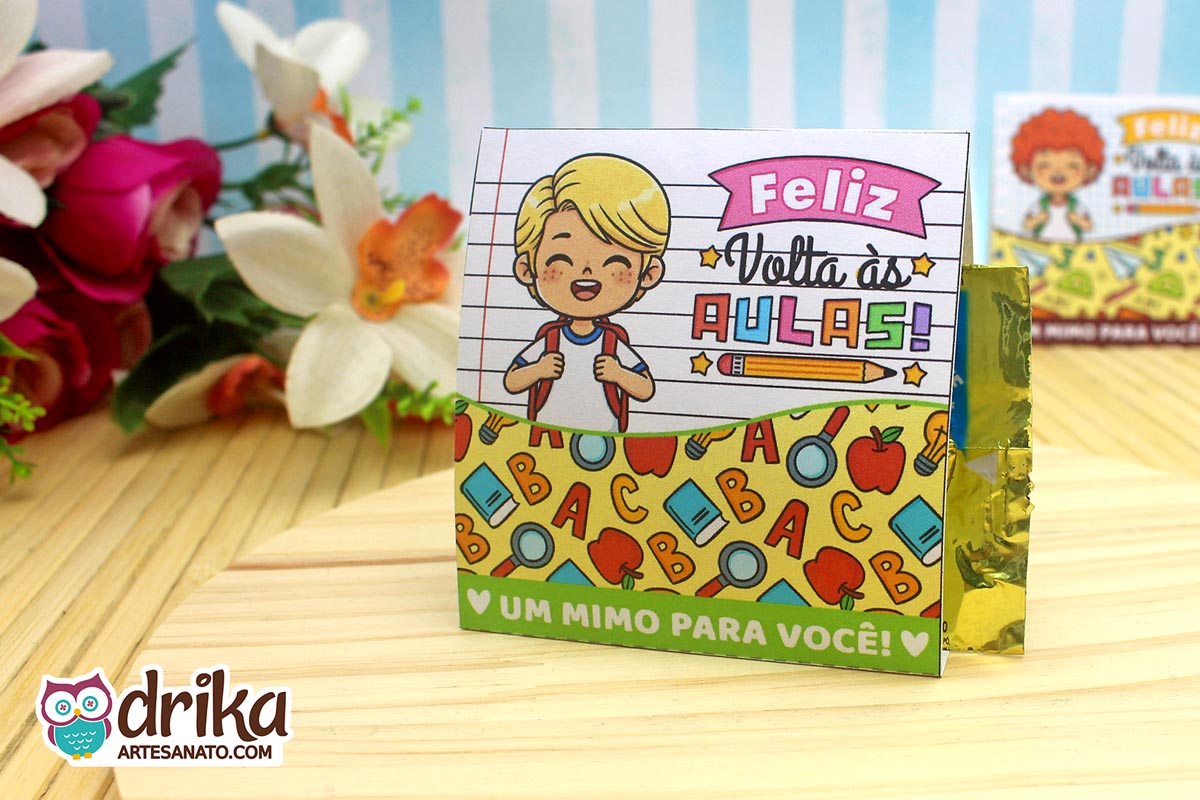 Porta-bombom de papel com menino loiro sorridente e mochila, em fundo de caderno. Decorado com letras, lupas e maçãs coloridas