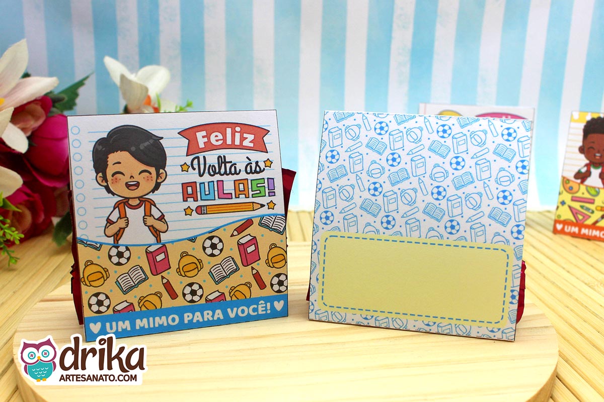 Porta-bombom de papel com menino de cabelo preto na frente e verso azul com estampa de material escolar e espaço para mensagem