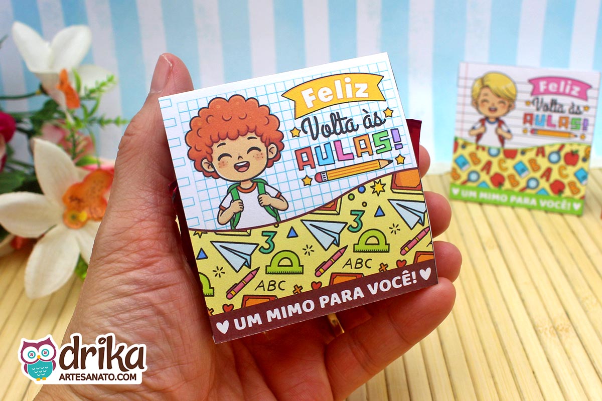 Mão segurando porta-bombom de papel com menino ruivo sorridente e estampa de material escolar em fundo de caderno