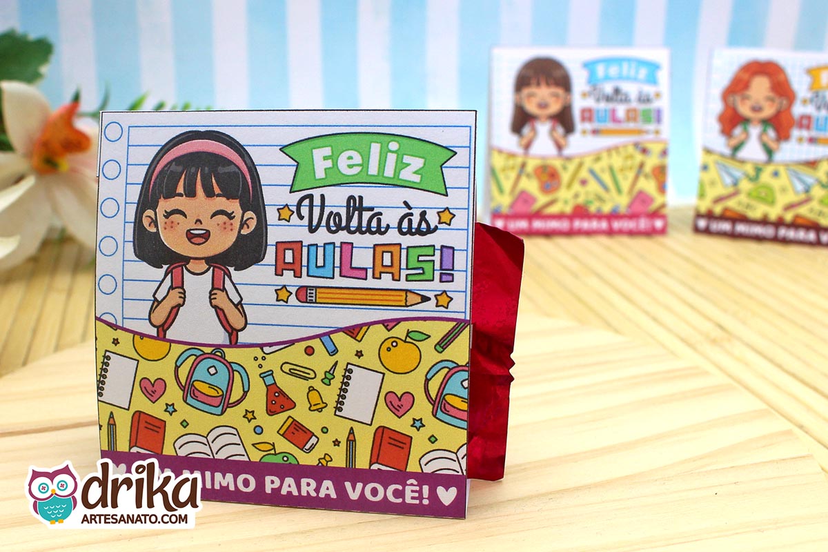 Porta-bombom de papel com menina de cabelo preto liso, mochila e "Feliz Volta às Aulas" em fundo de caderno pautado, bombom vermelho