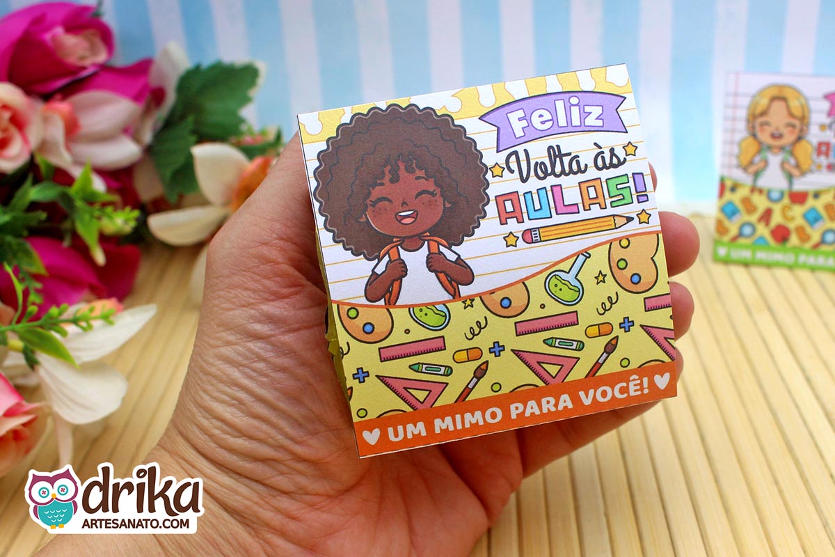 Mão segurando porta-bombom de papel com menina de cabelo crespo, decorado com tubos de ensaio e réguas