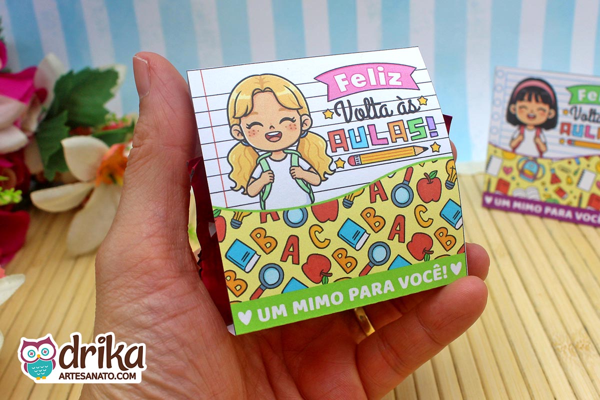 Porta-bombom de papel com menina loira de mochila e frase "Feliz Volta às Aulas", segurado por uma mão. Fundo pautado