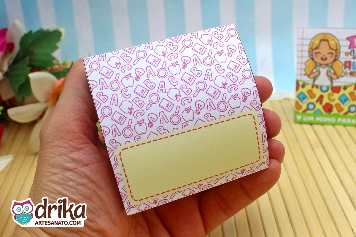 Verso de porta-bombom de papel com padrão de elementos escolares rosa e espaço para nome, segurado por uma mão