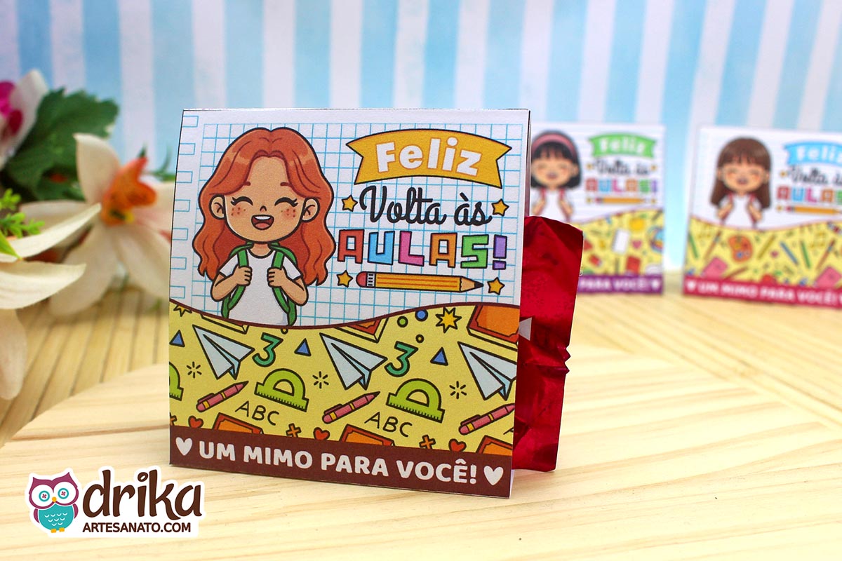 Porta-bombom de papel com menina ruiva, mochila e "Feliz Volta às Aulas" em fundo de caderno pautado, bombom vermelho