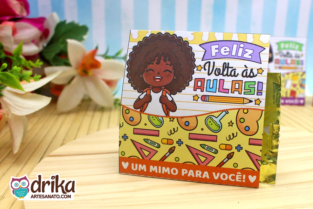 Porta-bombom de papel com menina de cabelo crespo, mochila e "Feliz Volta às Aulas" em fundo de caderno pautado, bombom dourado