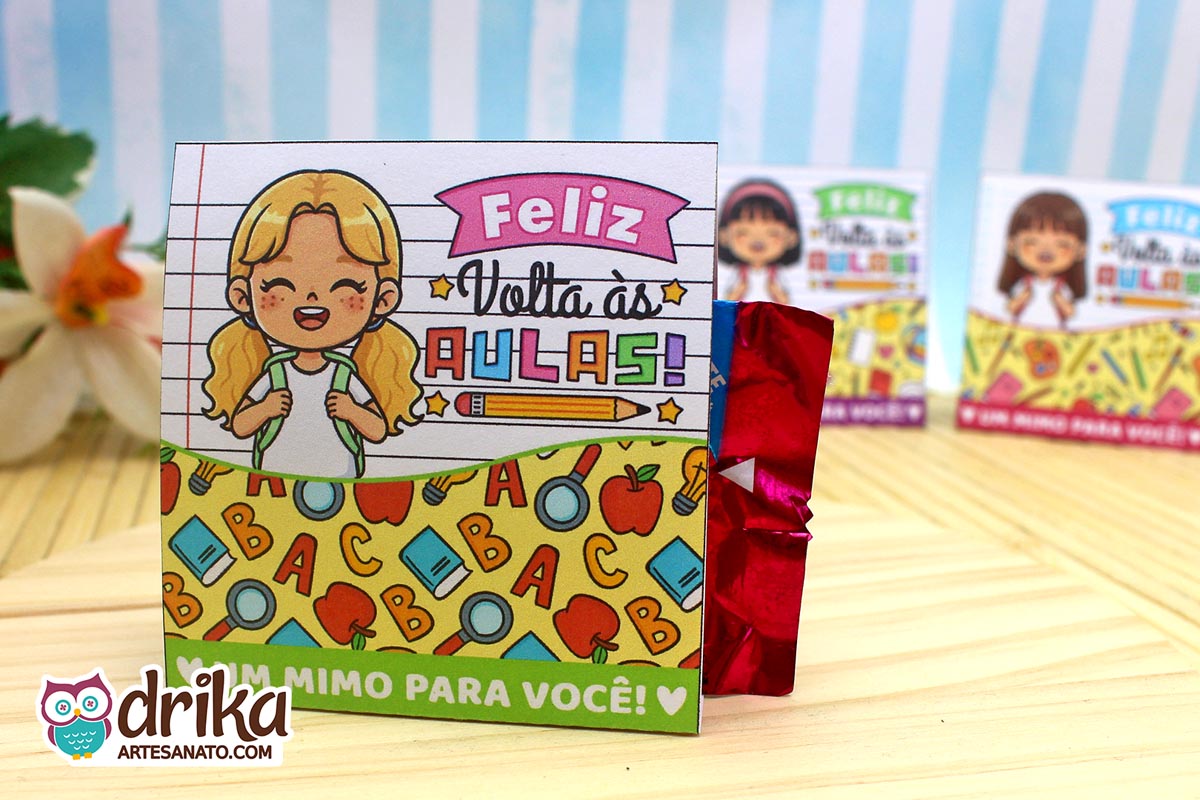 Porta-bombom de papel com menina loira, mochila e "Feliz Volta às Aulas" em fundo de caderno pautado, bombom vermelho