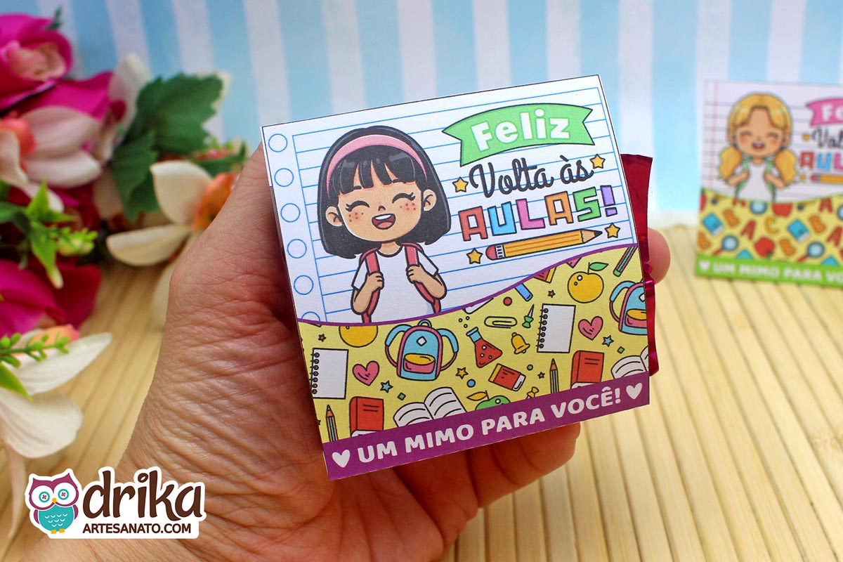 Mão segurando porta-bombom de papel com menina de cabelo preto liso e faixa, decorado com mochila e livros