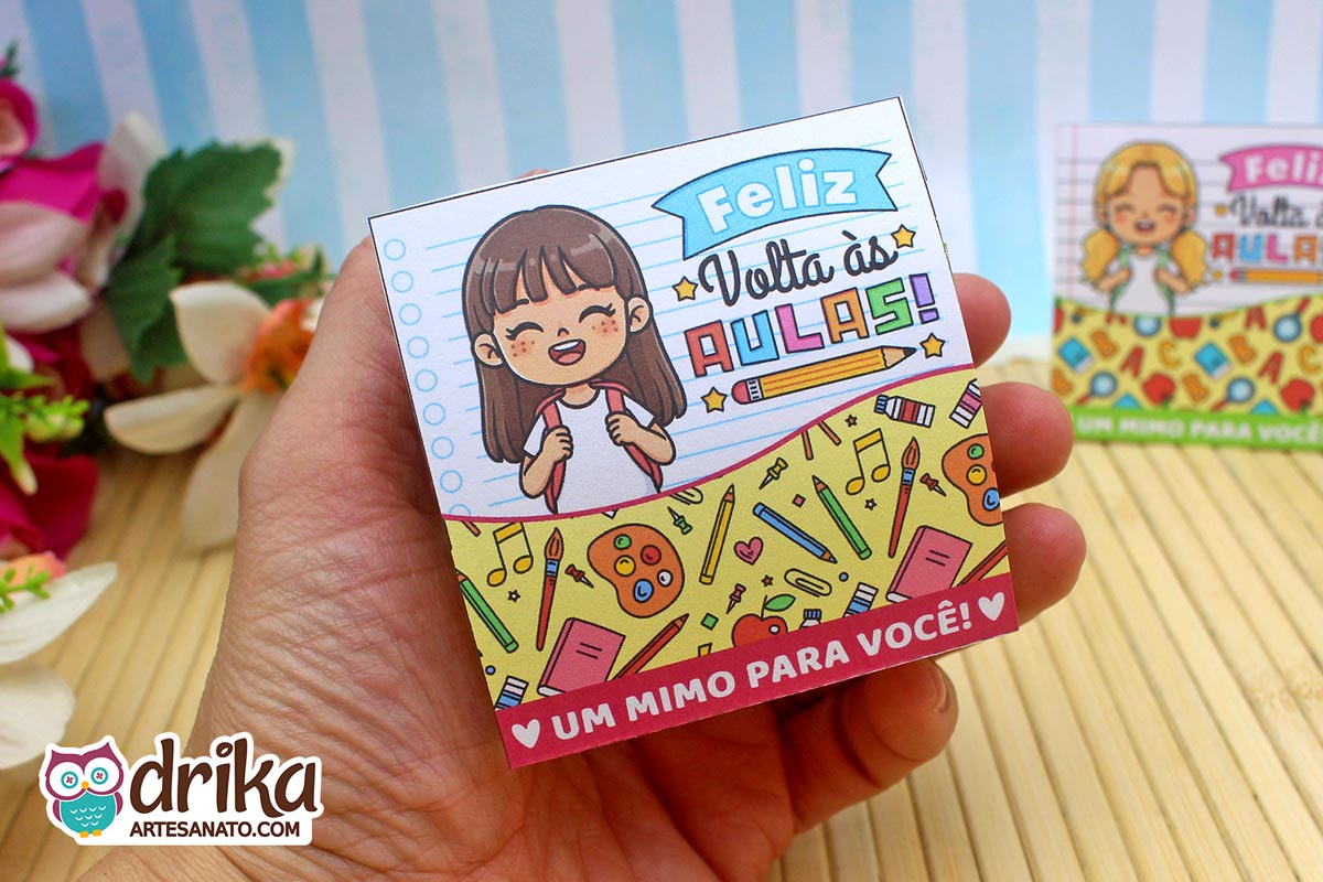 Mão segurando porta-bombom de papel com menina de cabelo castanho, decorado com paleta de tinta e pincéis