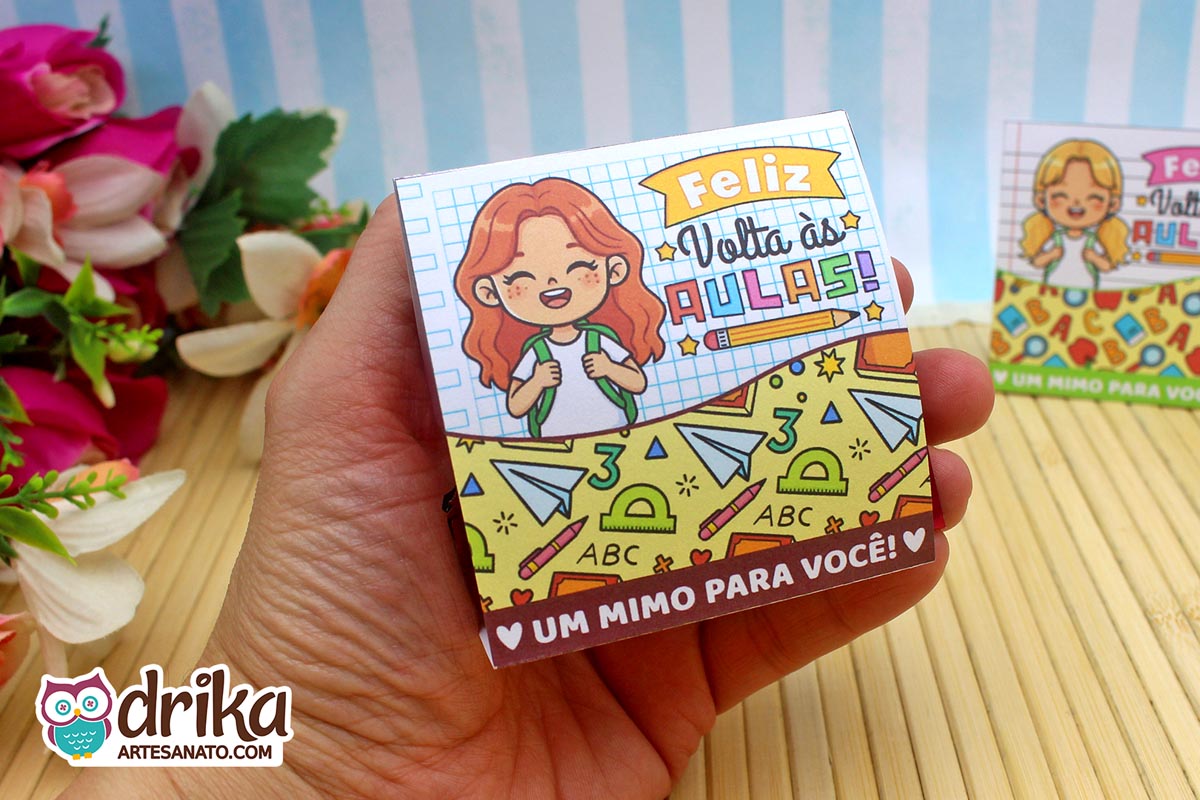 Mão segurando porta-bombom de papel com menina ruiva sorridente, decorado com réguas e letras do alfabeto