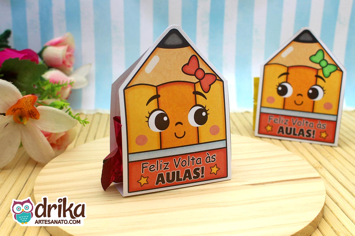 Dois porta-bombons de papel em formato de lápis com rostinhos de menina, um com laço vermelho e outro verde, sobre base de madeira