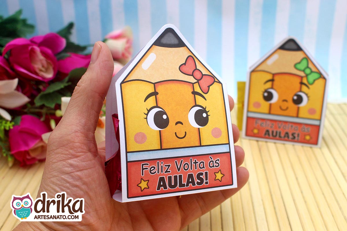 Mão segurando um porta-bombom lápis com rosto de menina e laço vermelho, mostrando o bombom na lateral