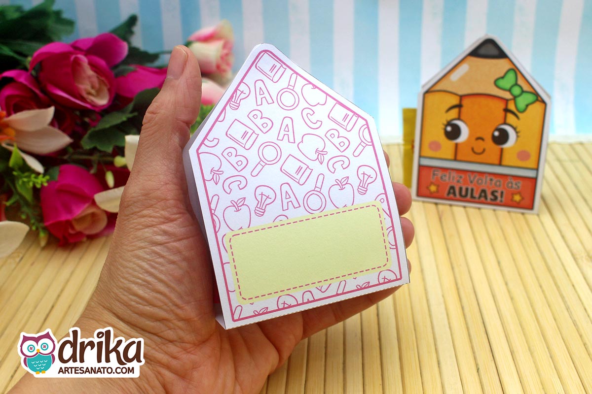 Mão segurando o verso de um porta-bombom lápis com estampa escolar e espaço retangular para escrever mensagem