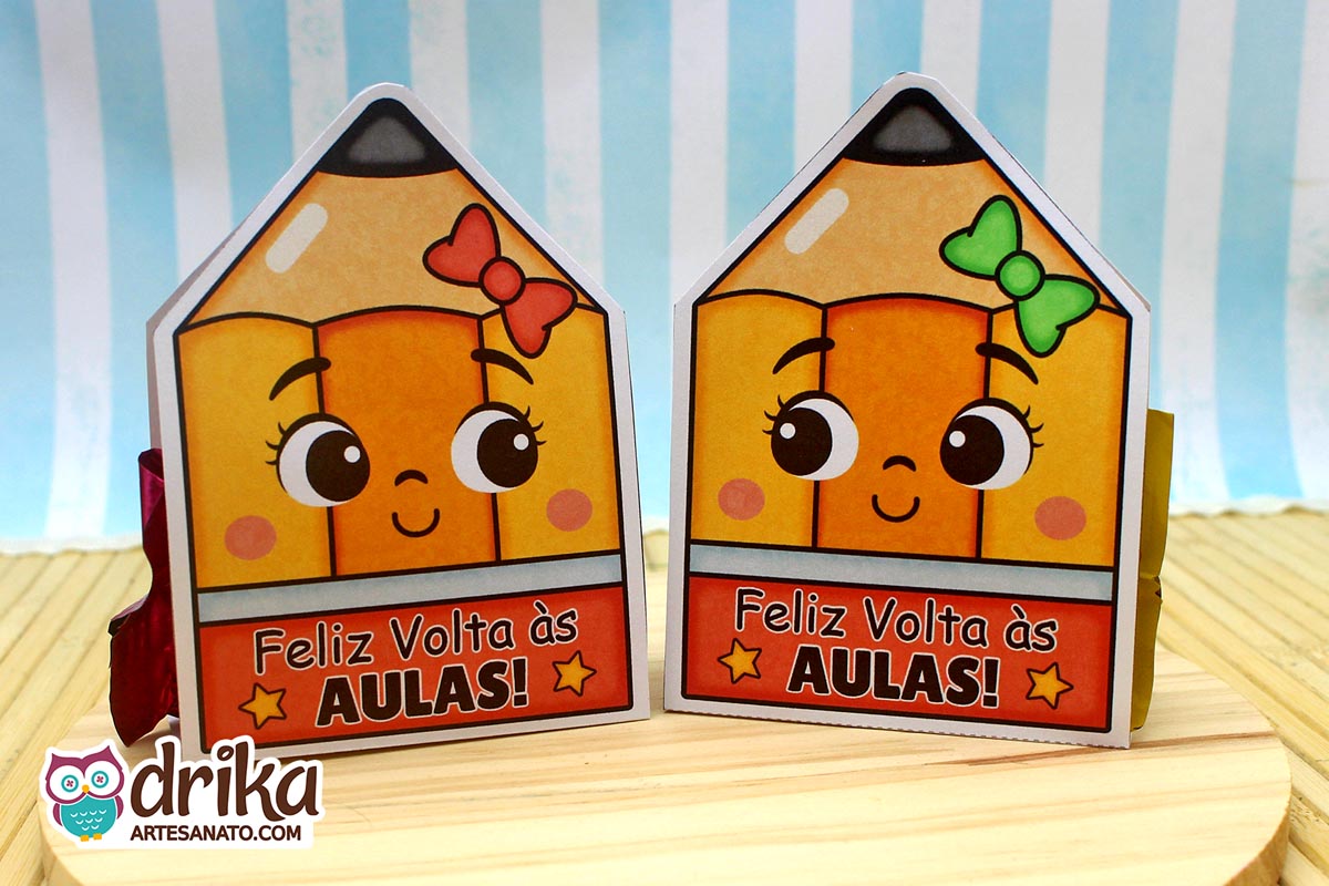 Dois porta-bombons de papel em formato de lápis com rostinhos de menina, um com laço vermelho e outro verde, lado a lado